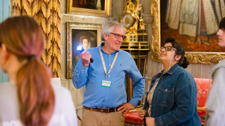 Visitors standing in a room that has paintings on the wall and gold coloured curtains. One person is holiding a torch, to show something to the other person. There is a ornate gold clock in the background.